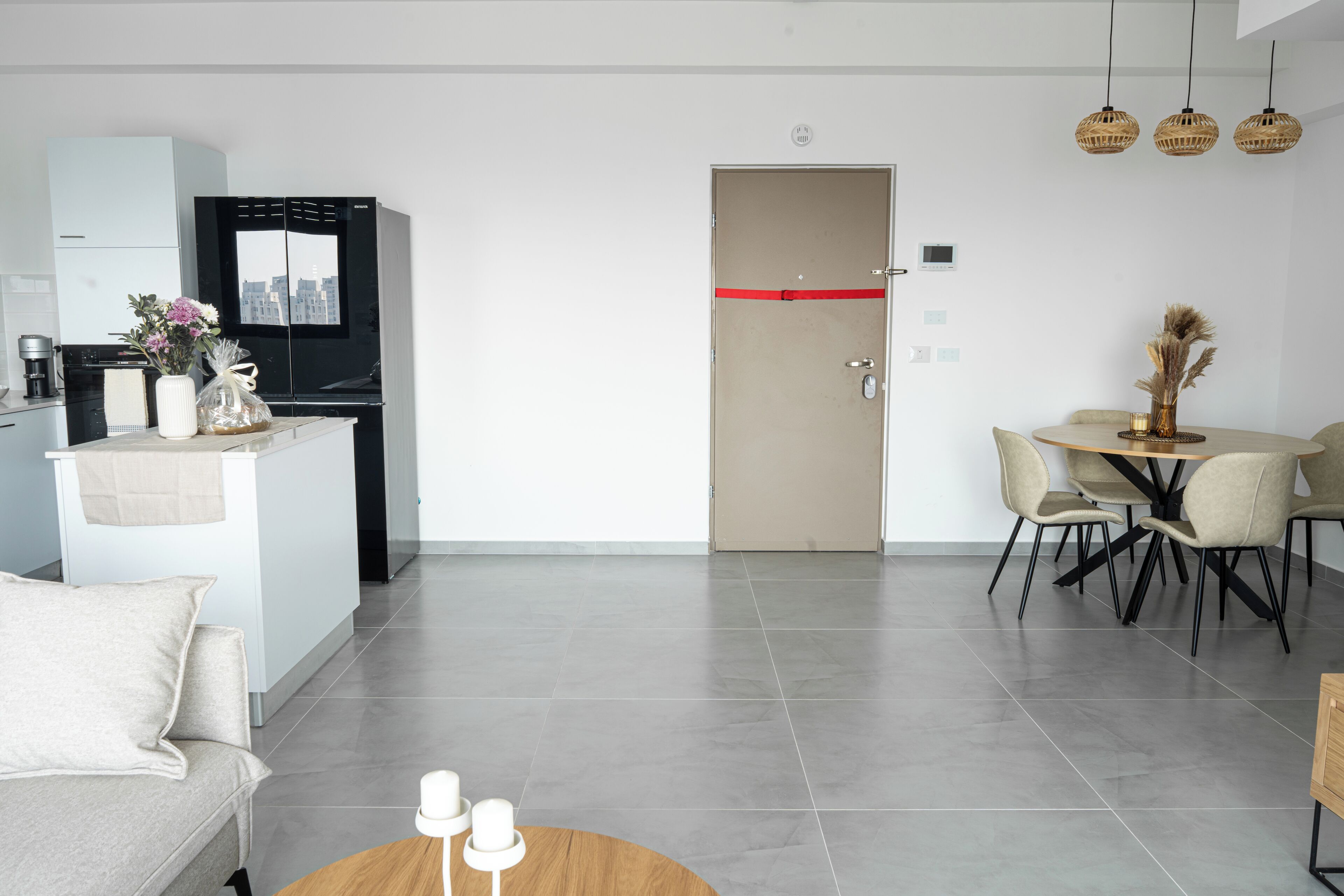 Un salon moderne comprenant un canapé, une table basse, un coin repas avec une table et des chaises, une cuisine avec un réfrigérateur, et une porte décorée d’un ruban rouge.