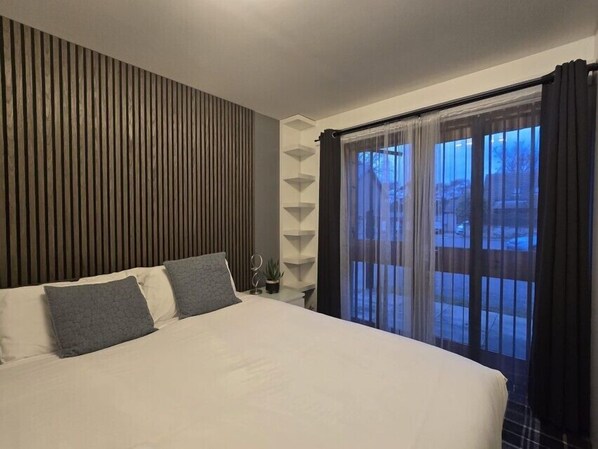 A modern hotel room with a large bed, a wooden slat wall, a small side table with a plant, and a view through sheer curtains. - Nairn Nights (Nairn)