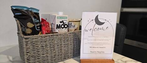 A welcome sign with a moon and stars design, a wicker basket with snacks and milk, and a kitchen in the background.