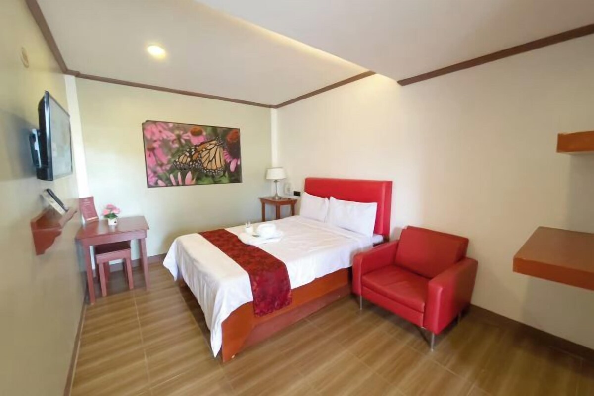Habitación de hotel con cama, una silla roja, un escritorio con lámpara y un cuadro en la pared.