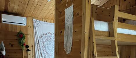 Interior de una cabaña de madera con literas, una escalera, un tapiz de macramé, un tapiz y una pequeña planta.