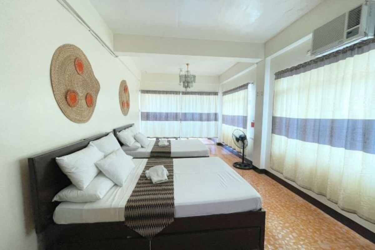 Habitación de hotel con una cama grande, una mesita de noche, un ventilador y vistas al exterior a través de cortinas translúcidas.
