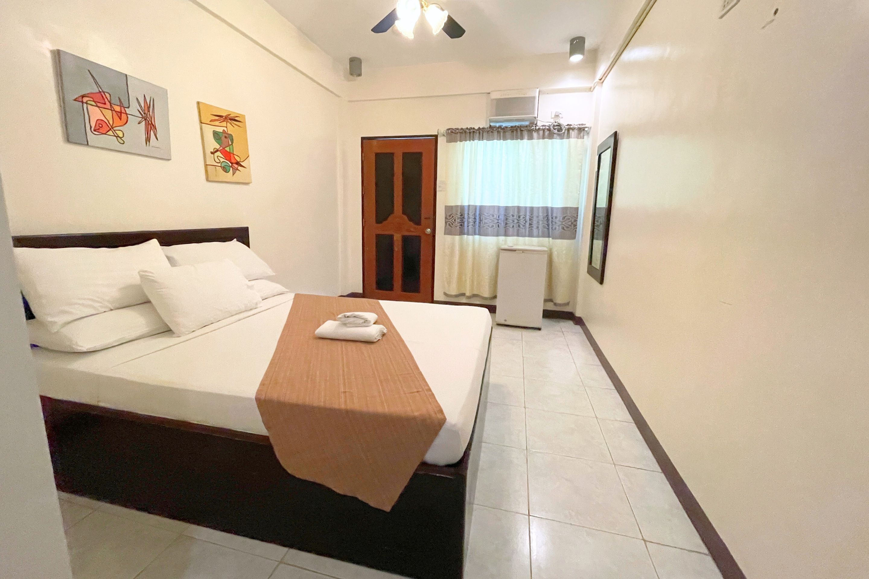 Habitación de hotel con cama, cabecera de madera, manta marrón, almohadas blancas, mesita de noche blanca, un cuadro en la pared y una ventana con cortinas.