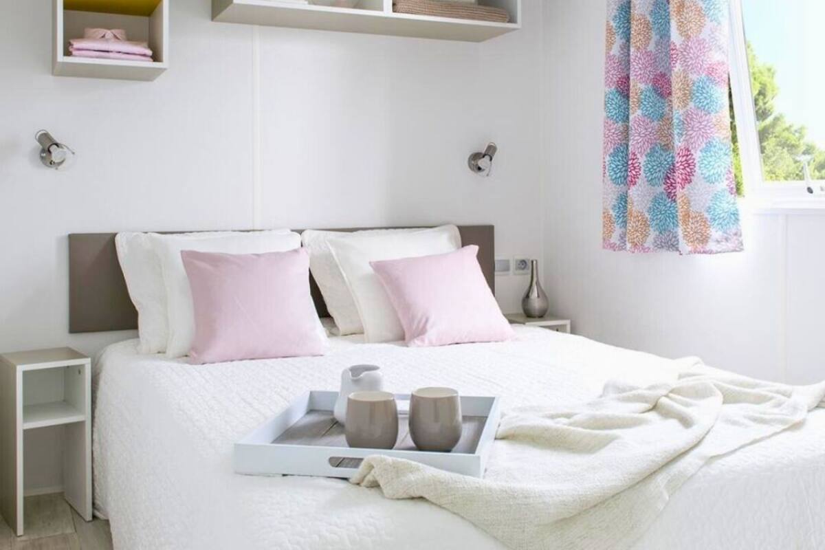 Una cama bien hecha con una bandeja de tazas y una jarrón, una mesita de noche y un ventanal con cortina colorida.