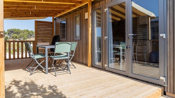 A covered outdoor area with a table and chairs, wooden deck, and sliding glass doors leading to an interior space. - Camping Pegomas (St.-Remy-de-Provence)