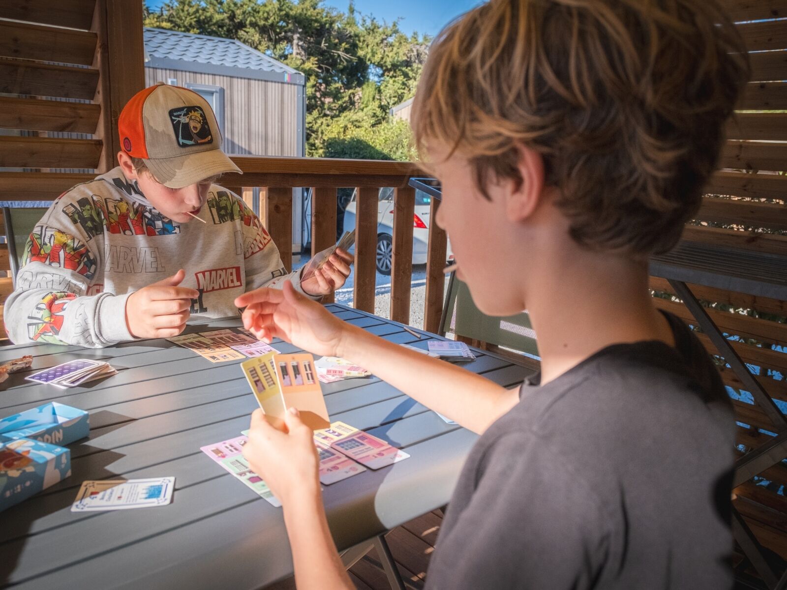 Two people playing a card game at an outdoor table with a wooden deck and a metal railing in the background.