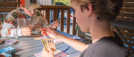 Two people playing a card game at an outdoor table with a wooden deck and a metal railing in the background.