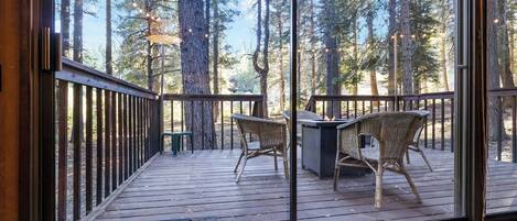 A wooden deck with wicker chairs and a table, overlooking a forest with tall trees.