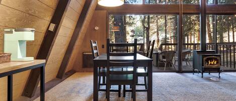 A cozy room with a dining table, chairs, a fireplace, and large windows offering a forest view.