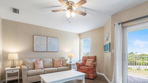 A living room with a beige sofa, two side tables, a coffee table, a patterned armchair, a ceiling fan, and a view of greenery through a sliding glass door. - Grand Caribbean 320 by Liquid Life (Orange Beach)