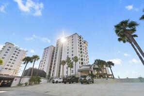 A parking area with several vehicles, including a white car and a black SUV, in front of a tall building with palm trees.