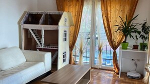 Ein modernes Wohnzimmer mit einem weißen Sofa, einem Holztisch und einem kleinen weißen Hausmodell. Vorhänge, ein Blumentopf und eine Deckenleuchte sind ebenfalls vorhanden.