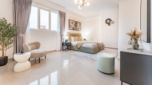 Een moderne hotelkamer met een bed, een zithoek met een fauteuil en een pouf, een klein bijzettafeltje, een potplant, een bureau met een vaas, en een grote spiegel.