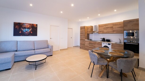 Ein modernes Apartment mit grauem Sofa, einem runden Esstisch mit gewebten Tischsets und einer Küche mit Holzschränken und Edelstahlgeräten.