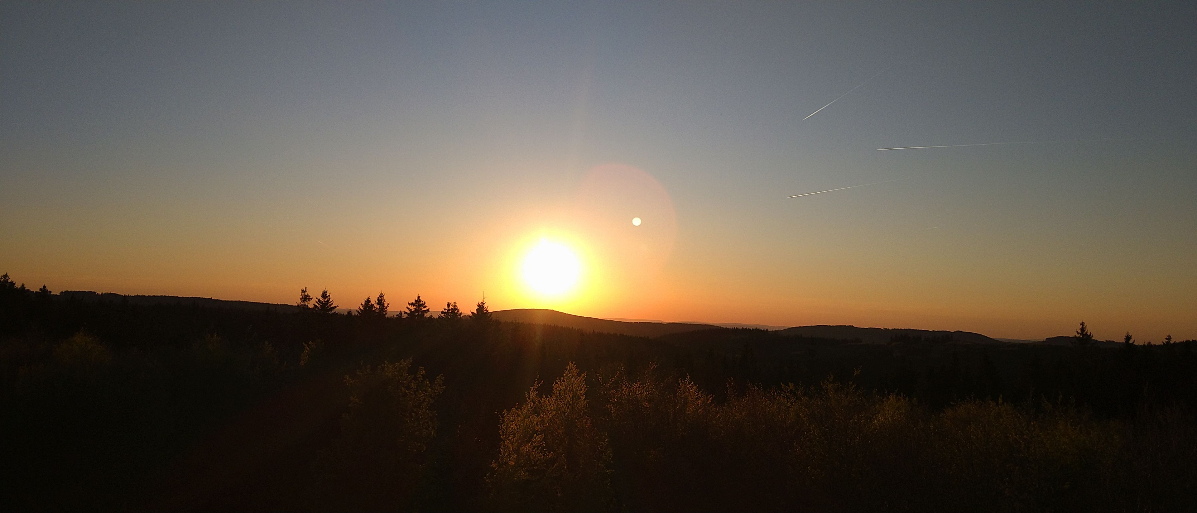 Um pôr do sol sobre uma paisagem acidentada, com árvores recortadas no céu.