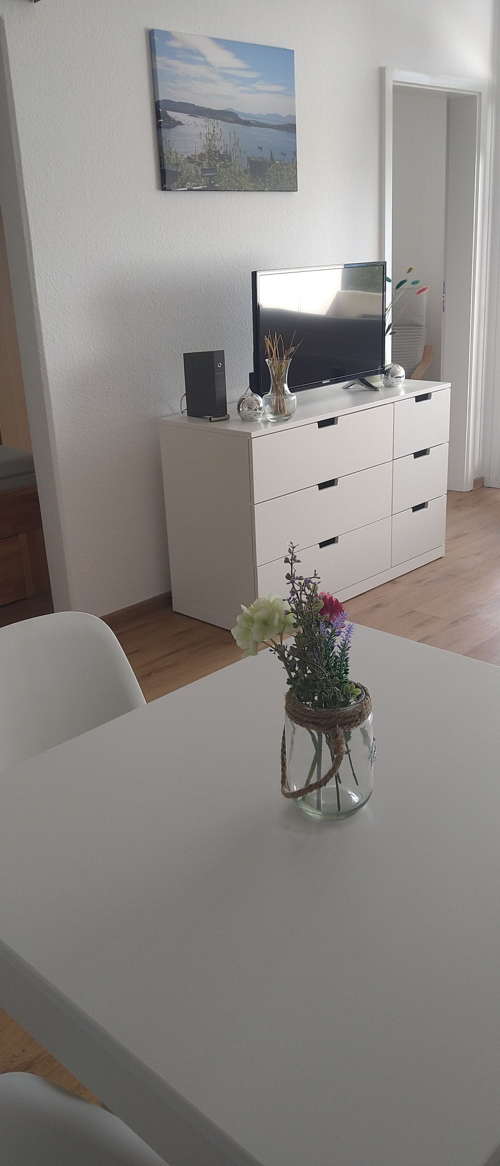 A room with a white table, a white cabinet, a television on top of the cabinet, and a painting on the wall.