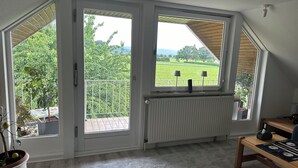 Quarto com uma janela grande com vista para uma paisagem verde, um radiador e uma mesa pequena com uma luminária.