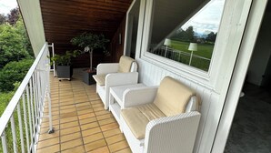 Varanda coberta com piso de azulejos, móveis de vime branco e vista para o verde.