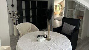 Uma área de refeições com uma mesa redonda coberta por toalha branca, cadeiras pretas e uma cadeira branca. Há uma estante preta na parede, um vaso com uma planta e uma pequena mesa com um liquidificador e um recipiente branco.