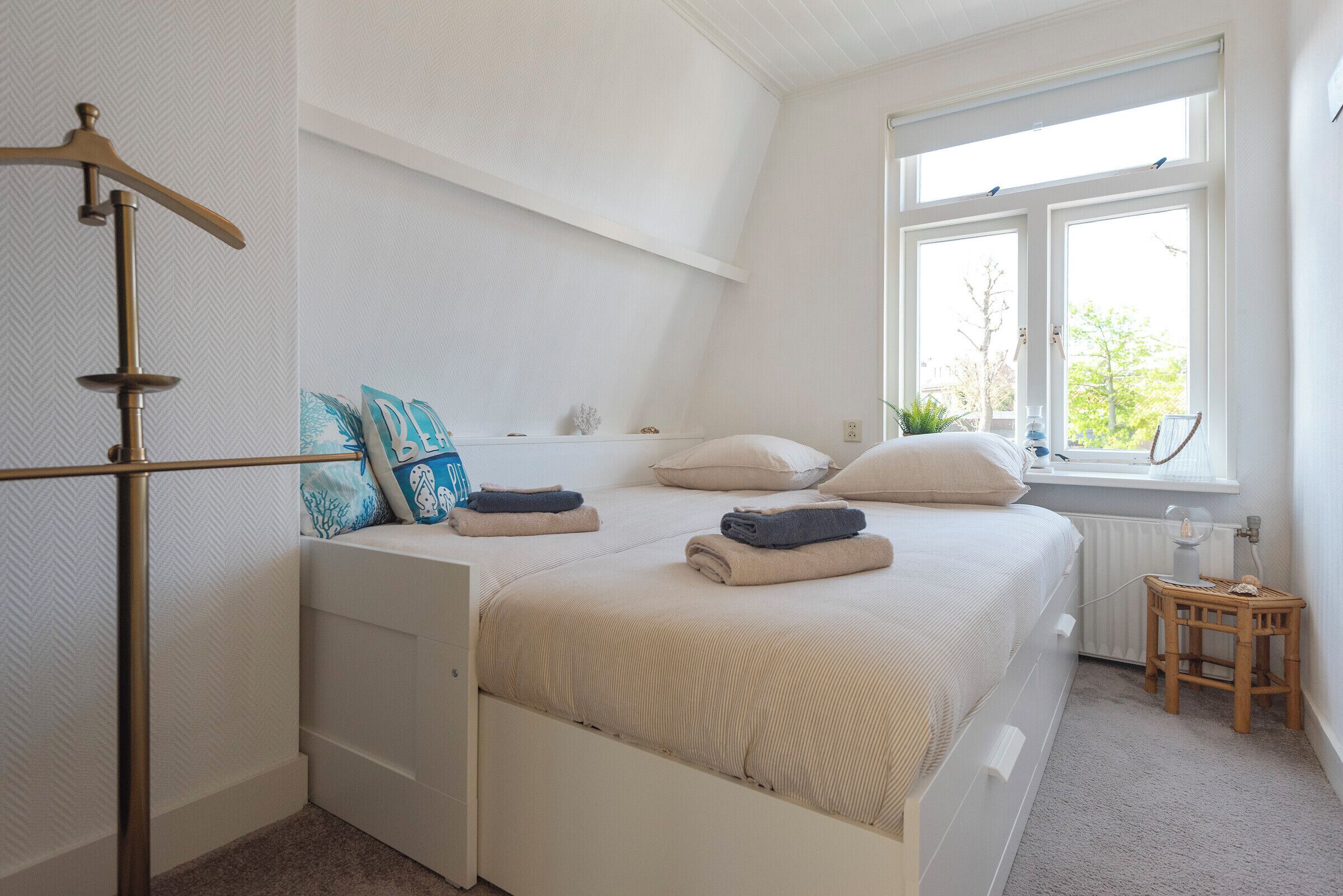 Ein Schlafzimmer mit Bett, Fenster, Heizkörper und einem kleinen Holzhocker.