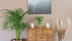 Et træskab med et billede i ramme over, en potteplante, en vase med blomster og en lysestage.