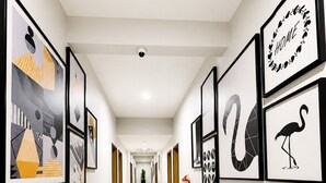 A hallway with framed artwork on the walls, a fire extinguisher, and a ceiling-mounted security camera. - Ixoras Hotel Lemba (Kinshasa)