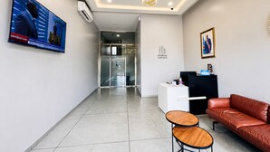A hotel reception area with a modern design, including a reception desk, a leather sofa, a small wooden table, and a television displaying a Zoom conference. - Ixoras Hotel Lemba (Kinshasa)