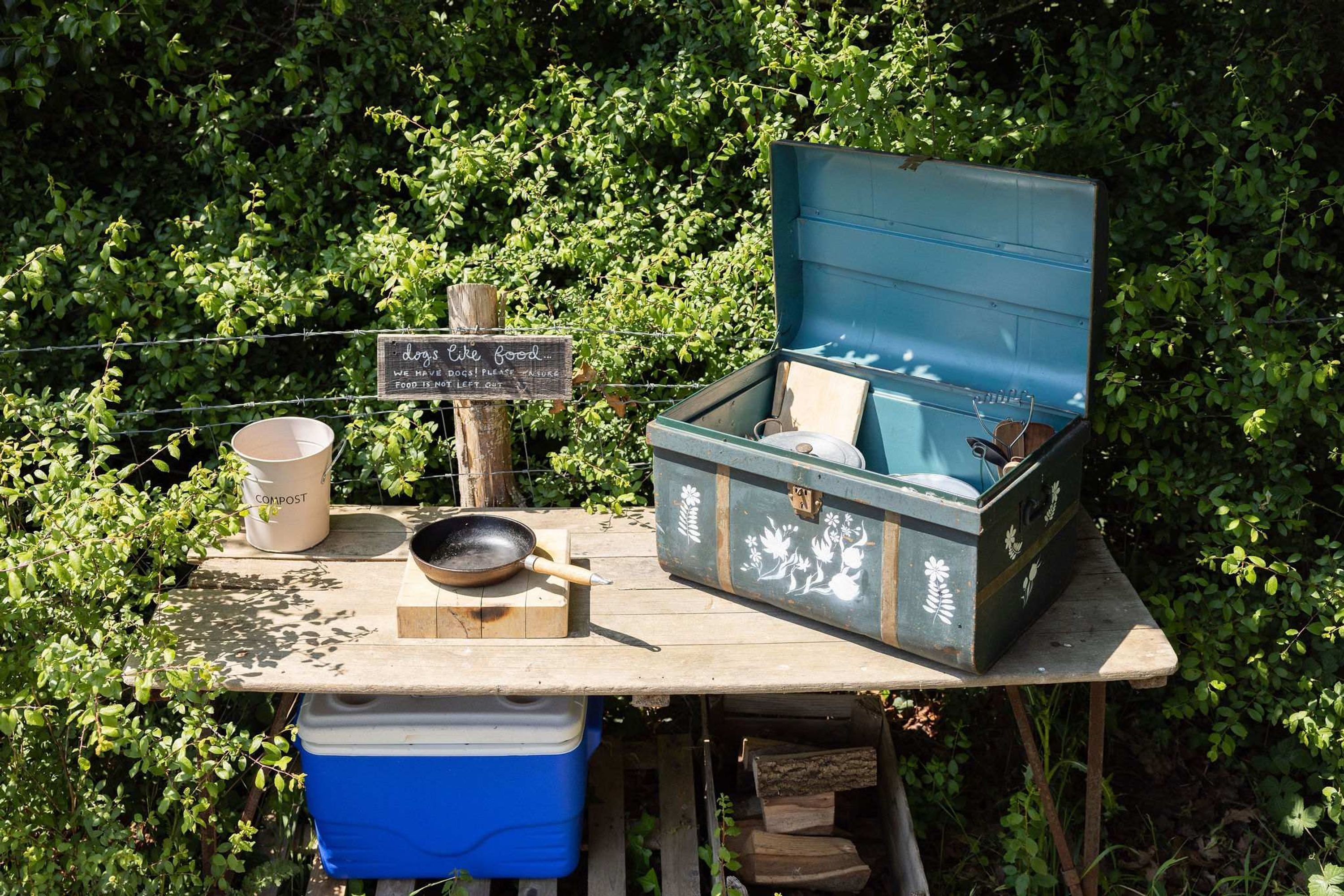 Un décor extérieur rustique comprenant une table en bois, un seau à compost, une glacière bleue, une poêle en métal et un coffre décoratif.