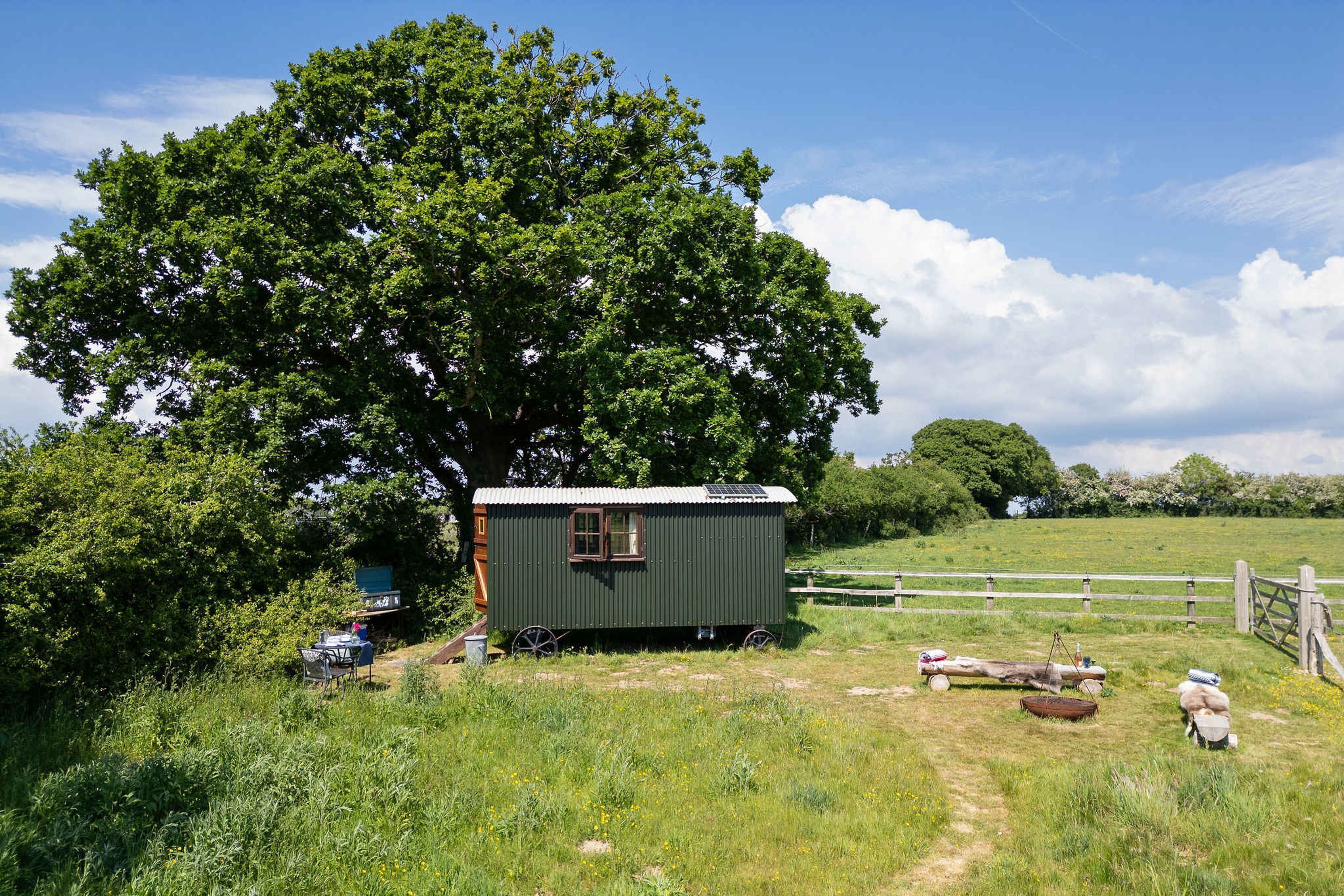 A green mobile home with a window and wheels, surrounded by a grassy field and trees.
