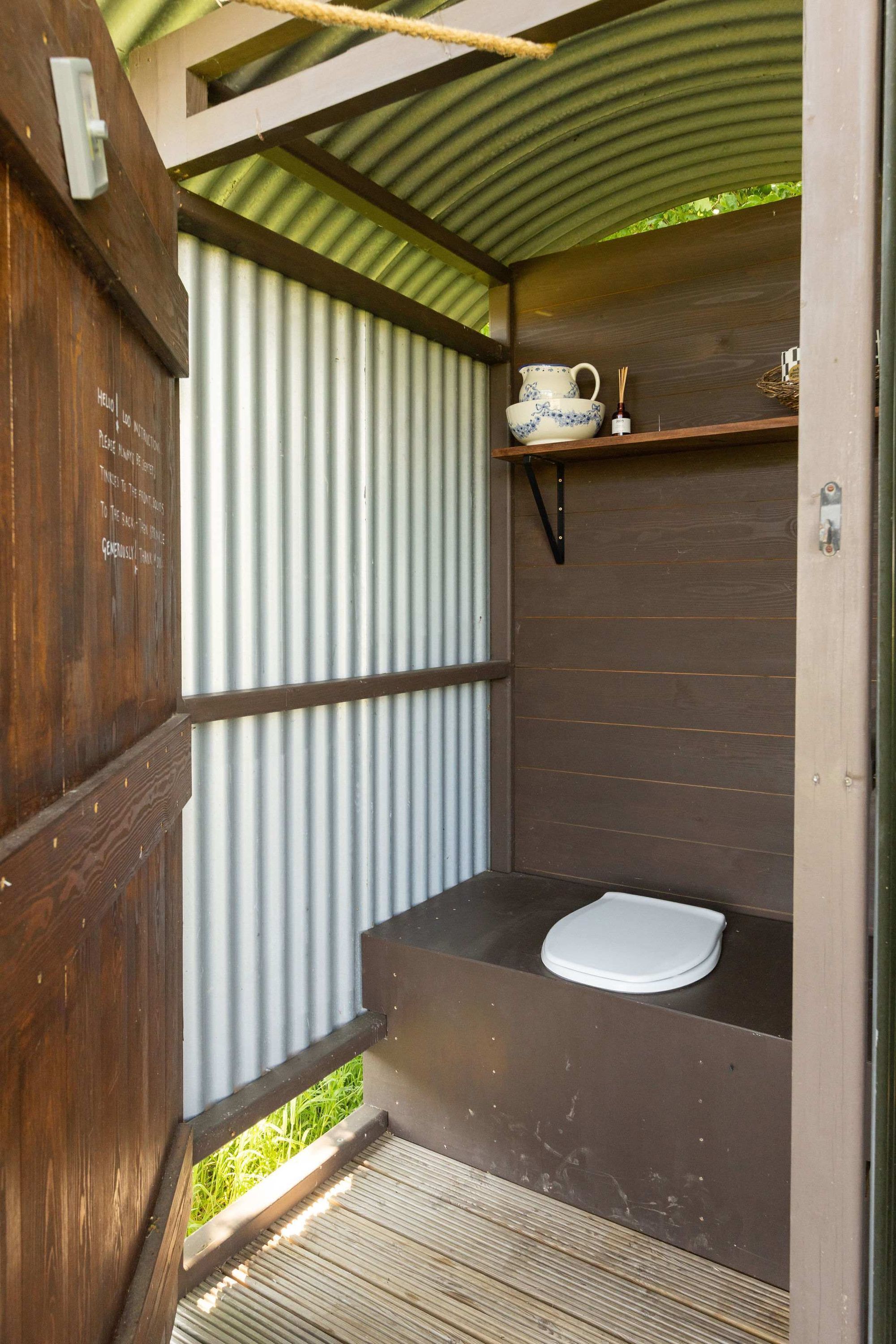 A compact, rustic bathroom with a wooden door, a corrugated metal wall, a small shelf with a cup and a candle, and a white toilet.