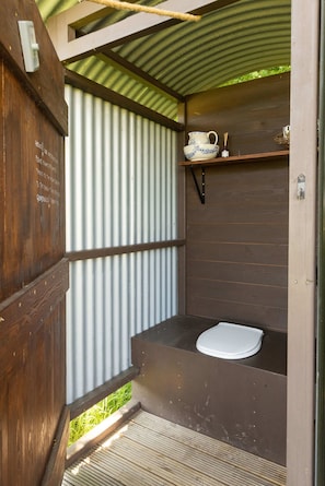 Um banheiro compacto e rústico, com porta de madeira, parede de chapa metálica, prateleira pequena com uma xícara e uma vela, e vaso sanitário branco.