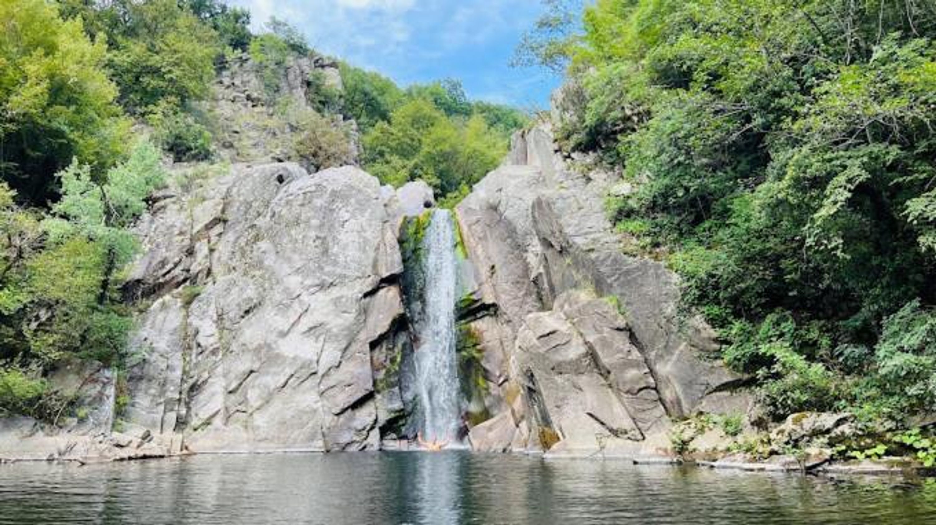 A waterfall cascading down a rocky cliff into a calm body of water, surrounded by lush greenery.
