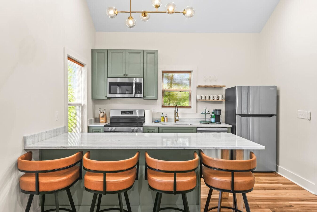 Uma cozinha moderna com ilha central, banquetas, geladeira de aço inoxidável e luminária pendente.