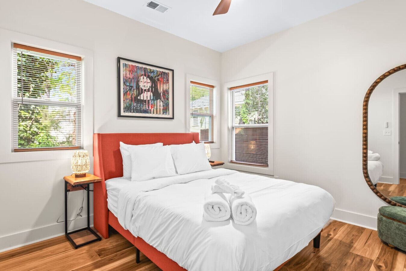 A modern bedroom with a large bed, white bedding, and a red headboard. There are two windows with blinds, a framed artwork above the bed, and a round mirror on the wall.