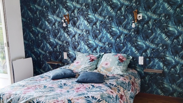 Una camera da letto con un letto, copripiumino floreale e carta da parati con motivo a foglia tropicale.