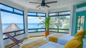 Een slaapkamer met twee bedden, grote ramen, een plafondventilator, een plant in pot en uitzicht op de oceaan.