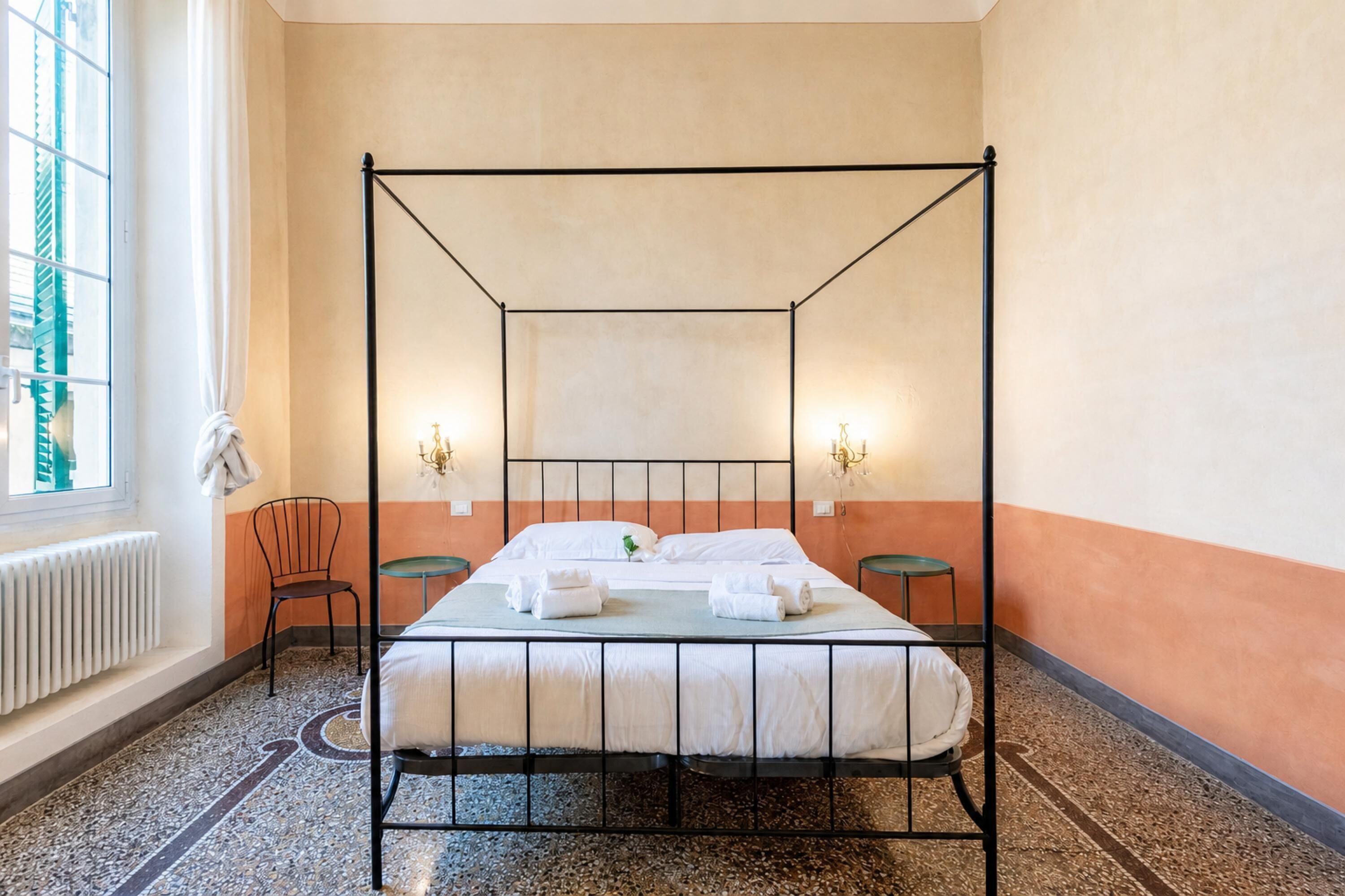 A four-poster bed with two bedside tables, a radiator, a chair, and a window with curtains.