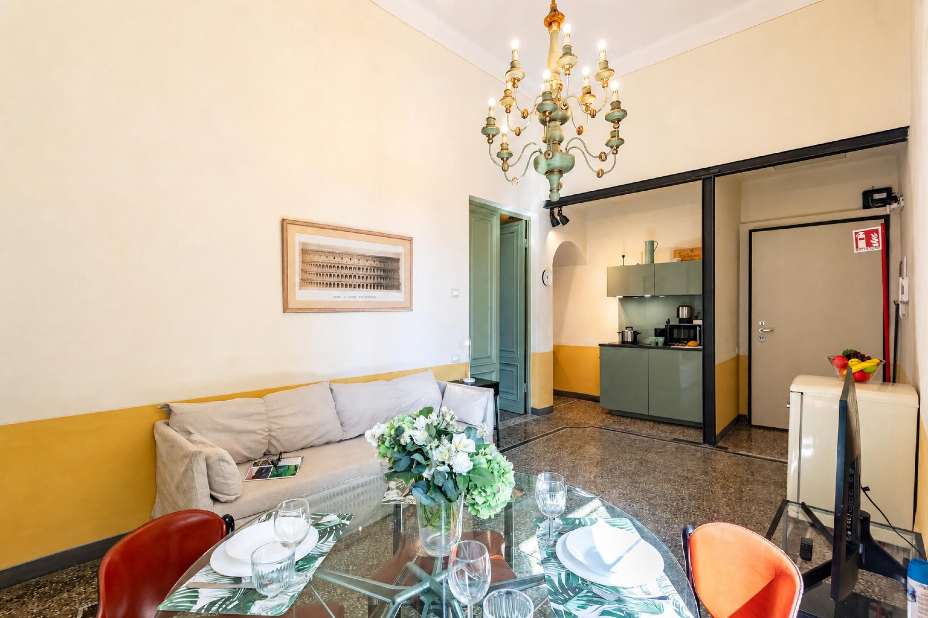 Un comedor con una mesa dispuesta para dos personas, un sofá, una araña y una cocina al fondo.