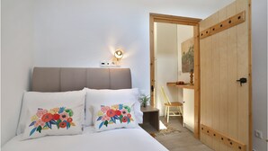A bedroom with a bed, a bedside table with a lamp, a wooden door, and a small desk with a chair.
