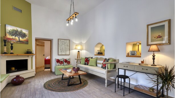 A living room with a sofa, coffee table, and a desk with a lamp. There is a fireplace, a painting, and a plant.