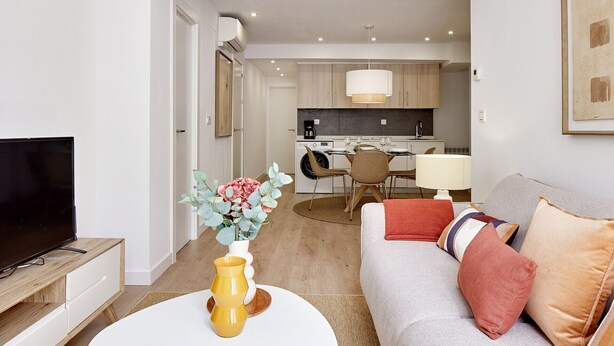 A modern living room with a flat-screen TV, a sofa with cushions, a coffee table with a vase, and a kitchen in the background. - Zenna | The Rentals Collection (San Sebastián)