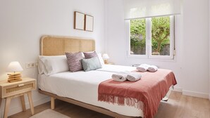 A neatly arranged bedroom with a bed, bedside tables, and a window with a view of greenery. - Zenna | The Rentals Collection (San Sebastián)