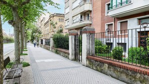 A sidewalk with a bench, a metal fence, and buildings with balconies and windows. - Zenna | The Rentals Collection (San Sebastián)