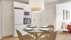 A modern kitchen with white cabinets, a built-in oven, and a microwave. There is a round glass table with four wicker chairs and a hanging light fixture above. - Zenna | The Rentals Collection (San Sebastián)