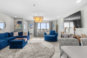 A modern living room with a sectional sofa, a blue armchair, and a grey armchair. There is a dining area with a table and chairs, and a large window providing a view. - T2201 at The Myrtle Beach Resort (Myrtle Beach)