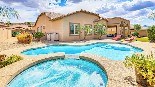 A swimming pool with a curved design, surrounded by a patio area with lounge chairs and palm trees. - Backyard Oasis w/ Fire Pit: Laveen Village Retreat (Laveen)