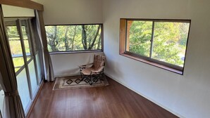 A room with a wooden floor, a folding chair, a patterned rug, and two windows with a view of greenery. - A mountain house for forest bathing in a hidden and unexplored part of Yakushima (屋久島町)