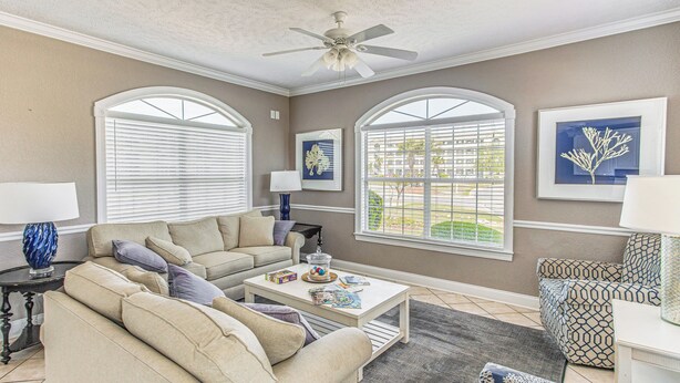 A living room with a beige sofa, a coffee table, and a patterned armchair. There are two windows with white frames and a ceiling fan.