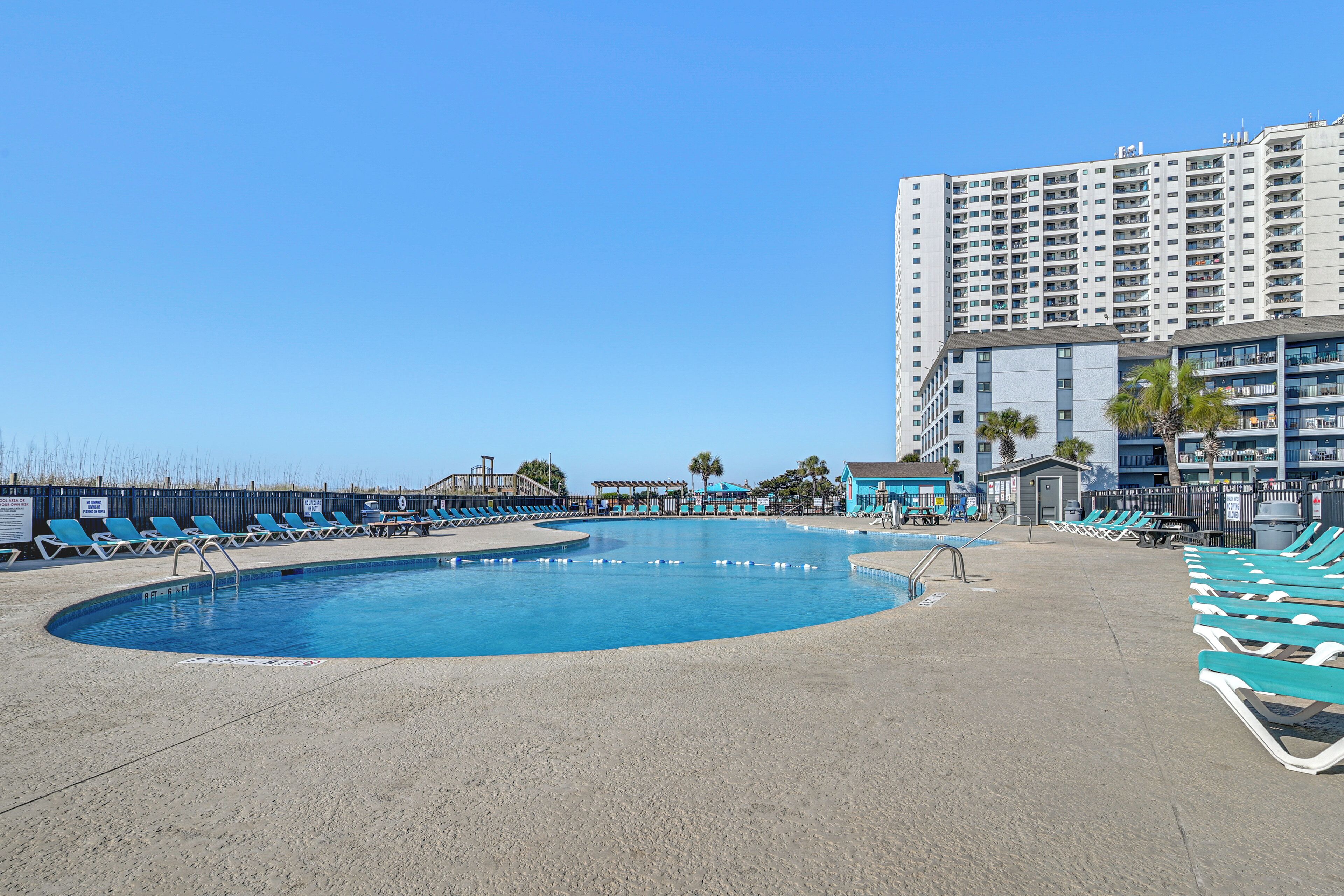 A pool area with lounge chairs, a building with multiple floors, and a clear sky.