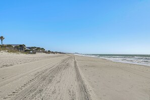 Ein sandiger Strand mit Spuren von Reifen, einer Palme und einem Gebäude im Hintergrund.